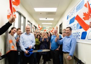 People at an office waving the Canadian flag. One person in front of the group cuts a ribbon with big scissors. 