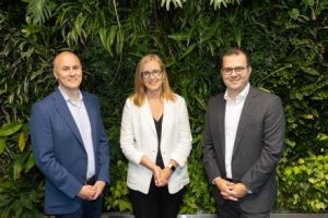 Three people formally standing in business suits in front of a wall of plants.