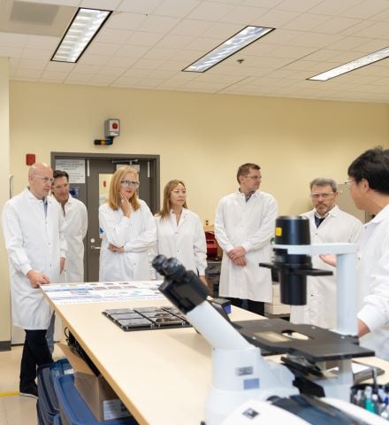People in white lab coats standing by microscopes in a lab.