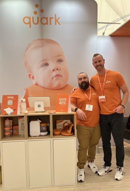 Quark Co-Founders, Senez and Gurinskas stand next to their products at a tradeshow booth. Baby photo in the back with their logo.