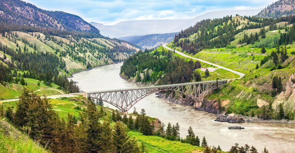 A scenic panoramic view of Williams Creek in the Cariboo region of British Columbia. It features a serene creek flowing through the landscape, with a charming bridge crossing over it.