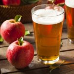 A glass of apple cider on a wooden surface beside fresh red apples and another glass, highlighting agricultural produce from British Columbia, Canada.