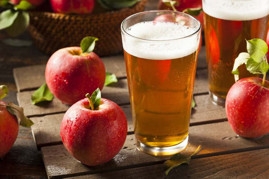 A glass of apple cider on a wooden surface beside fresh red apples and another glass, highlighting agricultural produce from British Columbia, Canada.