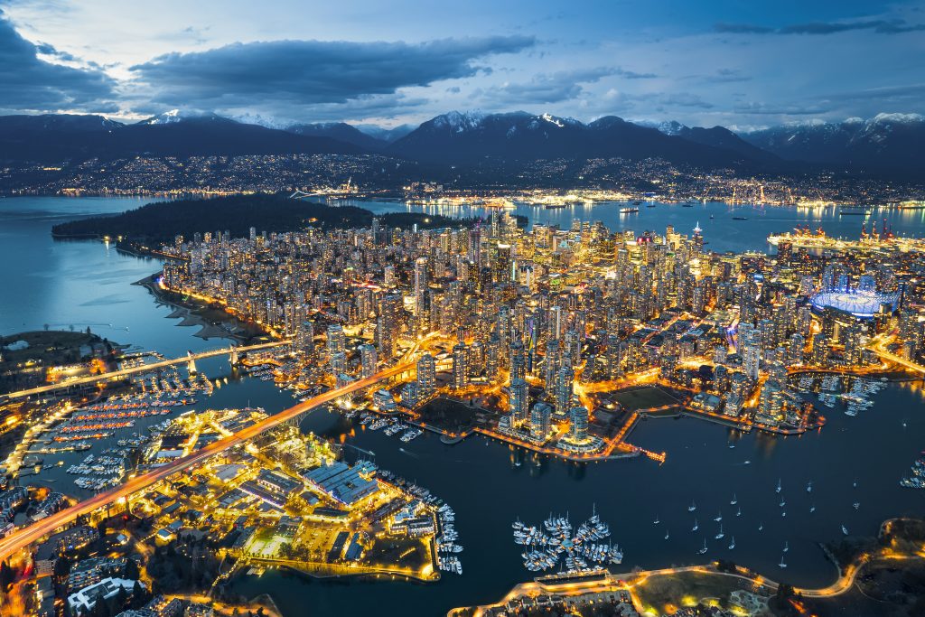 Aerial view of Vancouver, British Columbia at dusk, showing illuminated cityscape, port activity, waterways for trade, and nearby mountains.