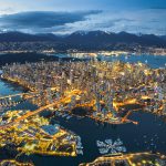 Aerial view of Vancouver, British Columbia at dusk, showing illuminated cityscape, port activity, waterways for trade, and nearby mountains.