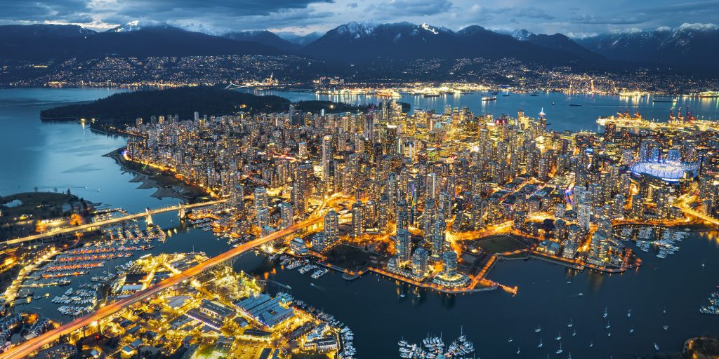 Aerial view of Vancouver, British Columbia at dusk, showing illuminated cityscape, port activity, waterways for trade, and nearby mountains.