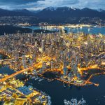 Aerial view of Vancouver, British Columbia at dusk, showing illuminated cityscape, port activity, waterways for trade, and nearby mountains.