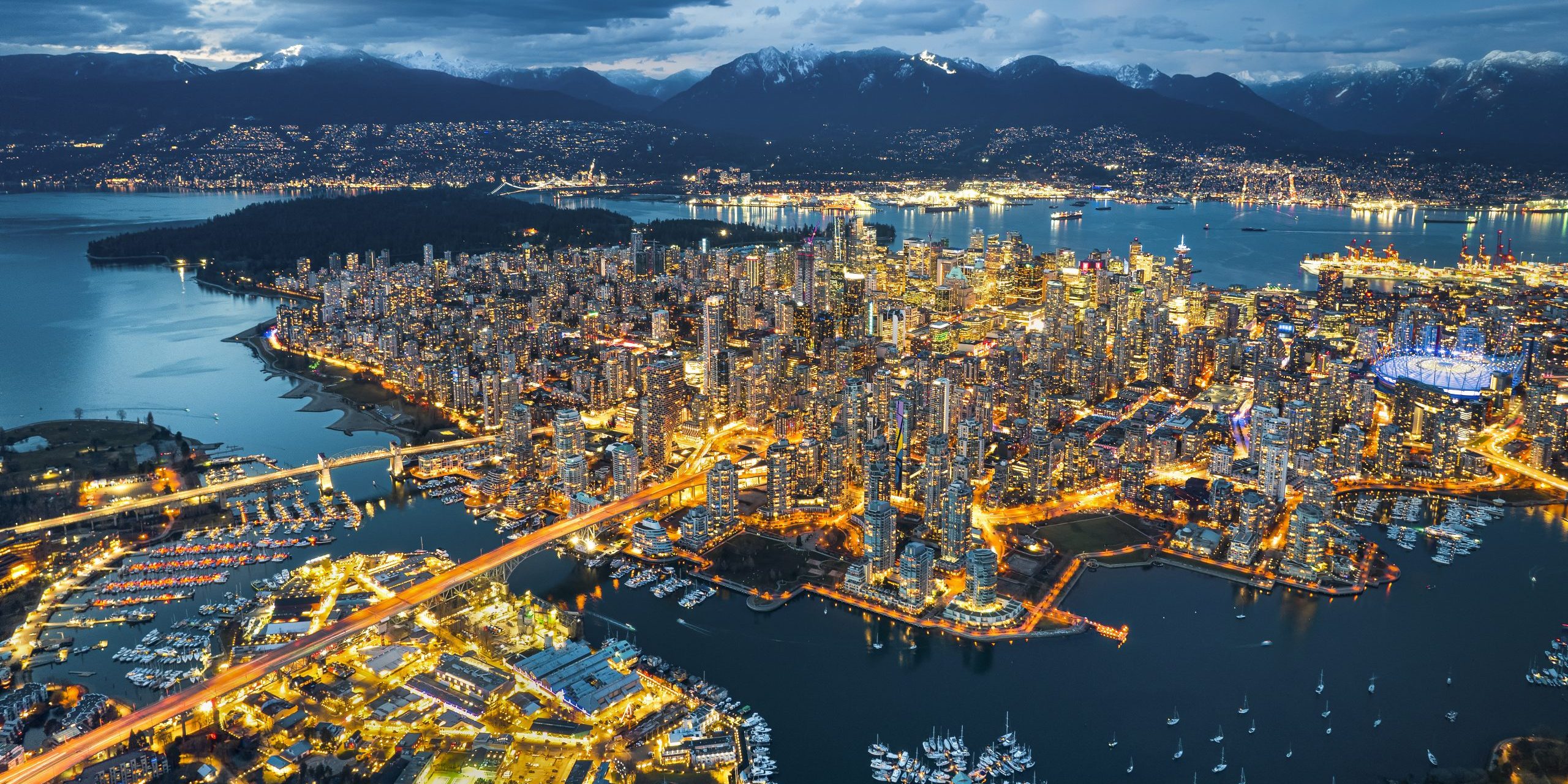 Aerial view of Vancouver, British Columbia at dusk, showing illuminated cityscape, port activity, waterways for trade, and nearby mountains.