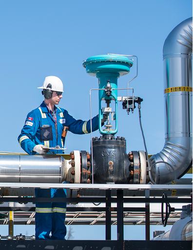A worker in safety gear inspects industrial piping and valves at an outdoor facility in British Columbia, Canada, under a clear blue sky.