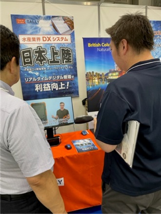 Two people with their backs to us are looking at a monitor at a trade show.